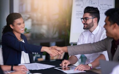 Three workers collaborating around desk, with a pair shaking hands, confirming their marketing ideas.